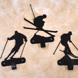 May include: Three black metal ski-themed wall hooks. Each hook features a silhouette of a skier in a different pose. The hooks are mounted on a light brown background.