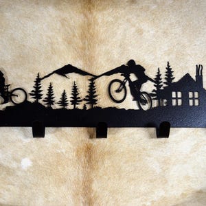 May include: Black metal key rack with a silhouette of two mountain bikers riding through a mountain landscape with a cabin in the background. The rack has five hooks.
