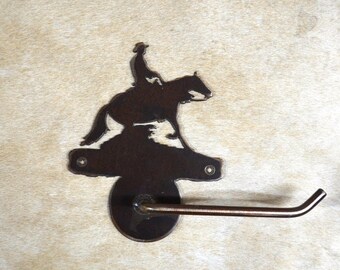 May include: A rustic metal toilet paper holder featuring a silhouette of a cowboy riding a horse. The holder is brown and has a simple design.