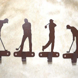 May include: Four metal golf-themed wall hooks. Each hook features a silhouette of a golfer in a different stance, with a hook below. The hooks are a dark brown color and are mounted on a light-colored surface.