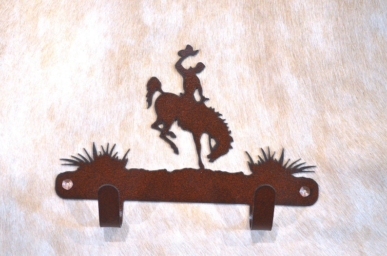 K&ouml;nnte beinhalten: Rustikaler Wandhaken aus Metall mit einer Silhouette eines Cowboys, der einen buckelnden Bronco reitet. Der Haken hat zwei Haken an der Unterseite und eine braune Rost-Optik.