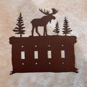 May include: Rustic brown metal light switch cover with a moose and pine trees silhouette. The cover has four switch openings.