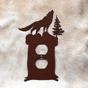 May include: A rusty brown metal wall plate with a wolf howling at the moon silhouette. The wolf is standing on a rock with a pine tree behind it. The plate has a standard electrical outlet cutout.