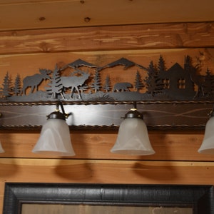 May include: A rustic metal light fixture featuring a silhouette of a woodland scene, including trees, mountains, a cabin, and animals. The fixture has five frosted glass light bulbs and a brown wooden base.
