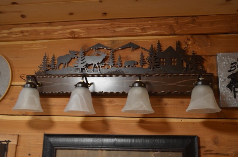 May include: A rustic metal light fixture featuring a silhouette of a woodland scene, including trees, mountains, a cabin, and animals. The fixture has five frosted glass light bulbs and a brown wooden base.