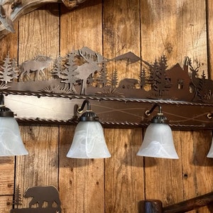 May include: A rustic metal light fixture with a cut-out design of a forest scene, including trees, mountains, and deer. The fixture has four frosted glass shades and is mounted on a wooden wall.