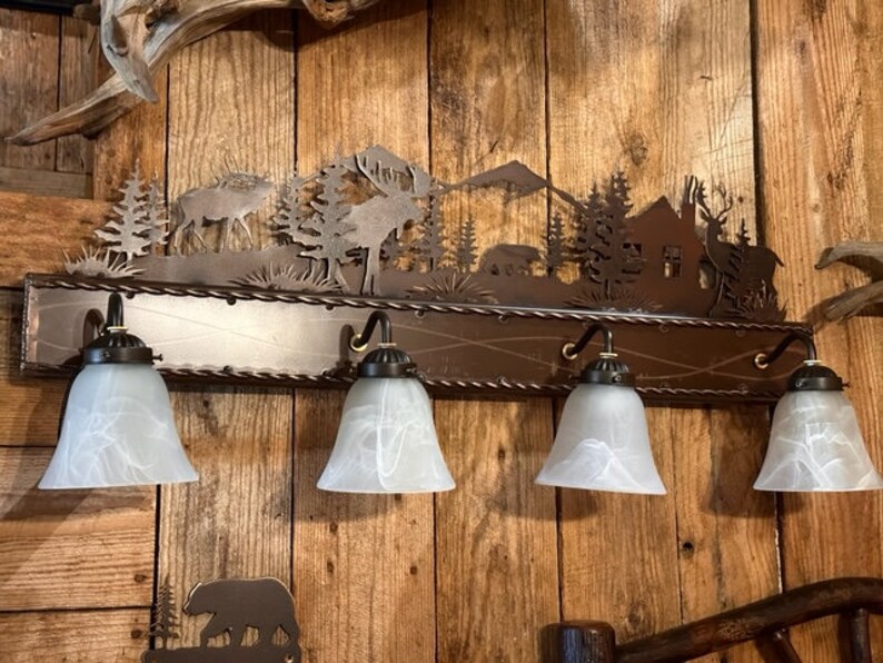 May include: A rustic metal light fixture with a cut-out design of a forest scene, including trees, mountains, and deer. The fixture has four frosted glass shades and is mounted on a wooden wall.