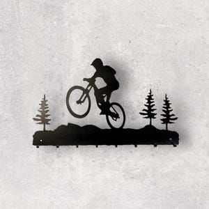 May include: Black metal wall art featuring a mountain biker mid-air, silhouetted against a backdrop of stylized trees and a mountain range. The art piece includes hooks for hanging items. The overall design is a nature-themed decorative item.