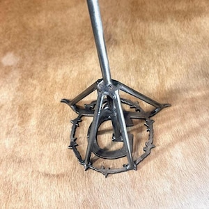 May include: A metal stand with a triangular base and a single upright rod. The base features a circular design with a stylized letter "G" in the center.