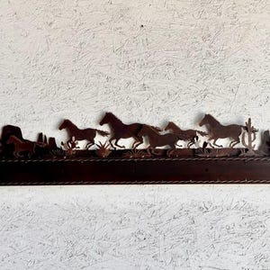 Pu&ograve; includere: Un decoro da parete in metallo con la silhouette di un branco di cavalli che corrono attraverso un paesaggio desertico. I cavalli sono marroni e il paesaggio &egrave; marrone chiaro. Il decoro &egrave; montato su uno scaffale in legno marrone scuro.