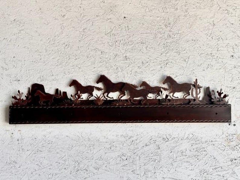 Pu&ograve; includere: Un decoro da parete in metallo con la silhouette di un branco di cavalli che corrono attraverso un paesaggio desertico. I cavalli sono marroni e il paesaggio &egrave; marrone chiaro. Il decoro &egrave; montato su uno scaffale in legno marrone scuro.