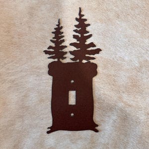 May include: A brown metal light switch cover with a silhouette of two pine trees. The cover is designed to fit a standard light switch.