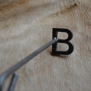 May include: A metal branding iron shaped like the letter "B" with a long handle. The branding iron is on a light brown surface.