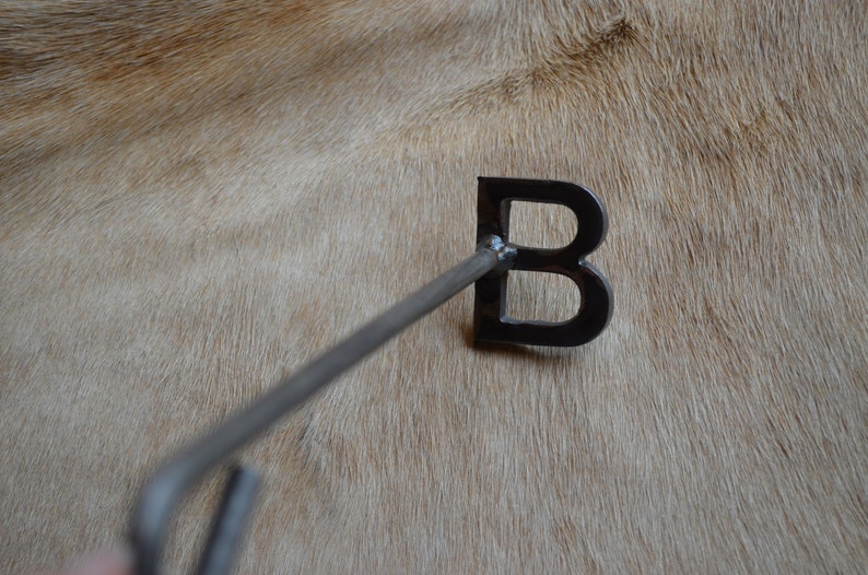 May include: A metal branding iron shaped like the letter "B" with a long handle. The branding iron is on a light brown surface.