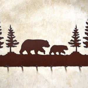 May include: Rustic metal key holder with a silhouette of two bears walking through a forest with five hooks. The key holder is painted in a rusty brown color.