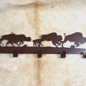May include: Metal wall mounted coat rack with five hooks. The rack is shaped like a silhouette of a herd of bison running. The rack is brown.
