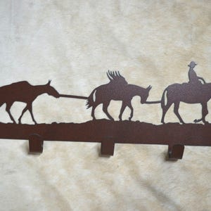 May include: A rustic metal wall hook featuring a silhouette of a cowboy riding a mule, followed by two other mules. The design includes two trees and four hooks. The color is a dark brown.