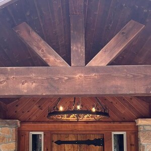May include: A wooden front porch with a dark brown stained wood ceiling and a large wooden beam. The ceiling has a decorative wood trim and a black metal chandelier with a circular design.
