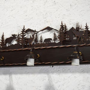 May include: A rustic metal light fixture with a woodland scene featuring trees, mountains, deer, and a bear. The fixture has three lights with clear glass shades and a dark brown wooden base.