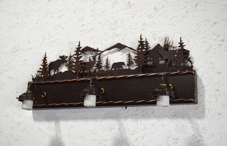 May include: A rustic metal light fixture with a woodland scene featuring trees, mountains, deer, and a bear. The fixture has three lights with clear glass shades and a dark brown wooden base.