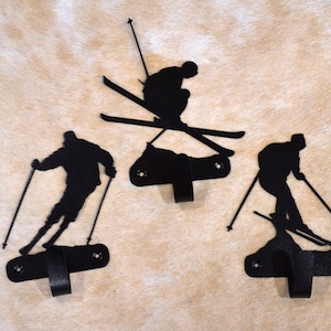 May include: Three black metal wall hooks shaped like skiers. Each hook features a silhouette of a skier in a ski pose with skis and ski poles. The hooks are mounted on a beige background.