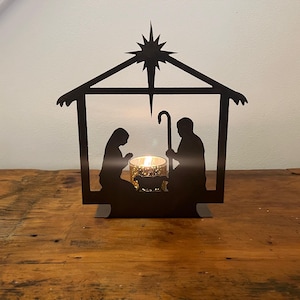 May include: A black metal nativity scene silhouette with a star on top. The scene includes figures of Mary, Joseph, and a shepherd, with a candle holder in the center. The candle holder contains a lit candle.