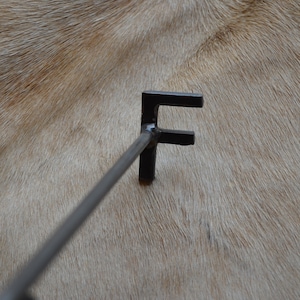 May include: A black metal branding iron with the letter "F" on the end. The branding iron is resting on a light brown surface.