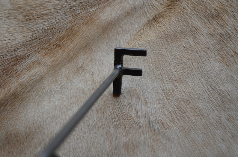 May include: A black metal branding iron with the letter "F" on the end. The branding iron is resting on a light brown surface.