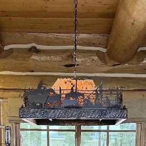 May include: A rectangular hanging pendant light fixture featuring a dark metal silhouette of a western rodeo scene with cowboys and horses. The fixture has a textured wooden frame base and is suspended from the ceiling by a metal chain link.