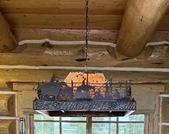 May include: A rectangular hanging pendant light fixture featuring a dark metal silhouette of a western rodeo scene with cowboys and horses. The fixture has a textured wooden frame base and is suspended from the ceiling by a metal chain link.