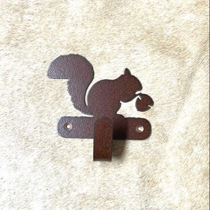 May include: A brown metal squirrel wall hook with a nut in its mouth. The hook is attached to a brown metal base with two screws.