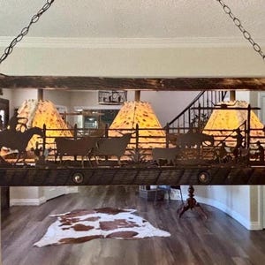 May include: A rustic, rectangular chandelier with a Western theme. The dark brown metal fixture features silhouettes of cowboys, cattle, and cacti. Three lampshades with a yellow and orange pattern are visible. The chandelier hangs from a chain.