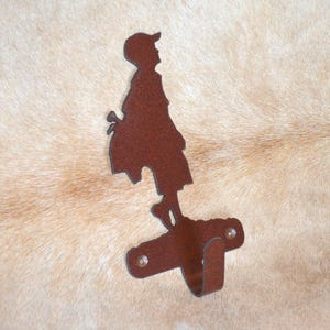 May include: A rusty brown metal silhouette of a person with a hat, used as a coat hook. The hook is attached to the bottom of the silhouette.