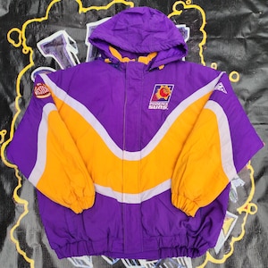 May include: A purple and gold Phoenix Suns jacket with a hood. The jacket features a large gold stripe across the chest and sleeves, with white accents. The Phoenix Suns logo is on the chest.