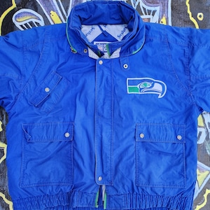 May include: A blue Seattle Seahawks jacket with a green and white logo on the chest. The jacket has a high collar, multiple pockets, and a zipper closure. The jacket is made of a lightweight material.