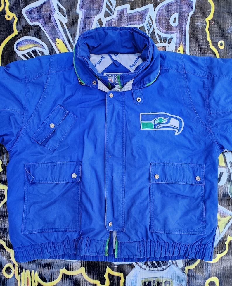 May include: A blue Seattle Seahawks jacket with a green and white logo on the chest. The jacket has a high collar, multiple pockets, and a zipper closure. The jacket is made of a lightweight material.
