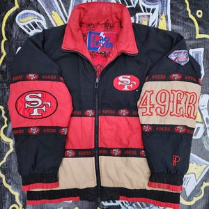 May include: A black, red, and tan Pro Layer NFL San Francisco 49ers zip-up jacket. The jacket features the 49ers logo on the chest and sleeves, with 49ERS text on the right sleeve, a red collar, and a black zipper. The interior lining has NFL branding.