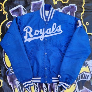 May include: A blue satin baseball jacket with white stripes on the collar and cuffs. The jacket has a white "Royals" logo on the front.