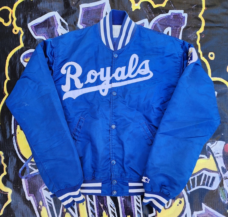 May include: A blue satin baseball jacket with white stripes on the collar and cuffs. The jacket has a white "Royals" logo on the front.