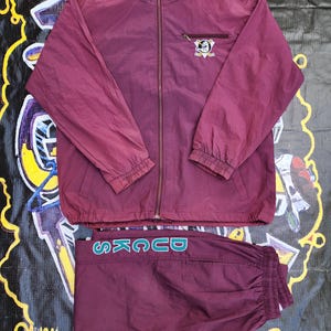 May include: A maroon tracksuit with a teal logo on the trousers and a teal and yellow logo on the jacket. The jacket has a zip fastening and a high collar.