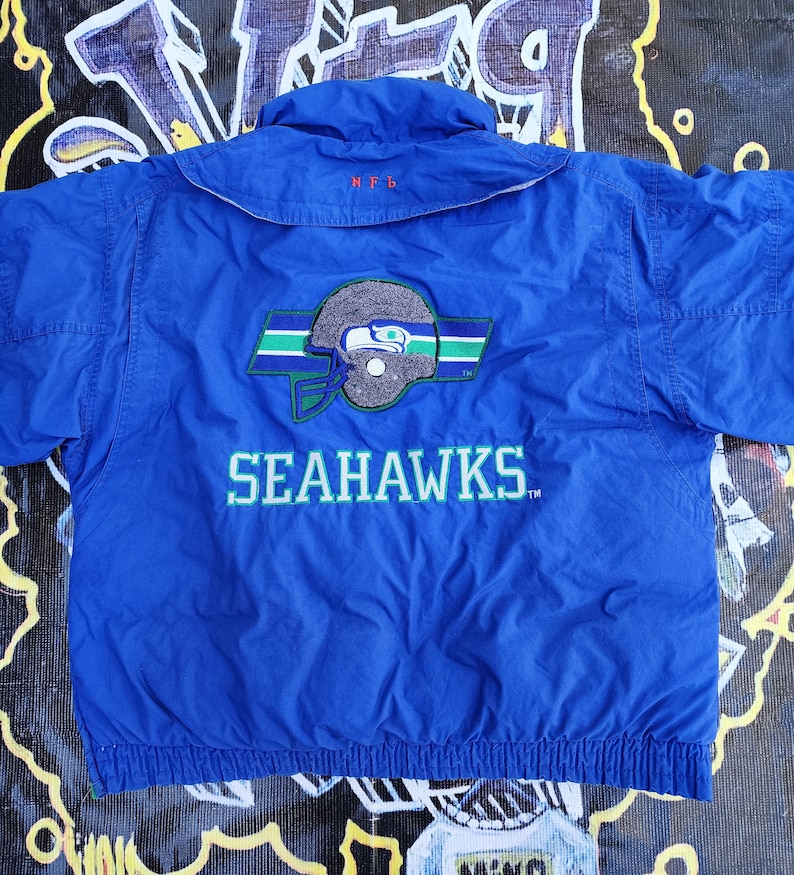 May include: A blue Seattle Seahawks jacket with the team logo and the word "SEAHAWKS" in white lettering. The jacket has a collar and a zipper. The NFL logo is embroidered on the collar.
