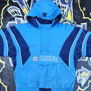 Puede incluir: Chaqueta con capucha azul claro y azul marino con la palabra "CAROLINA" y un logotipo en la parte delantera. La chaqueta tiene un bolsillo delantero y un logotipo de Starter en la capucha. La chaqueta est&aacute; hecha de un material brillante.