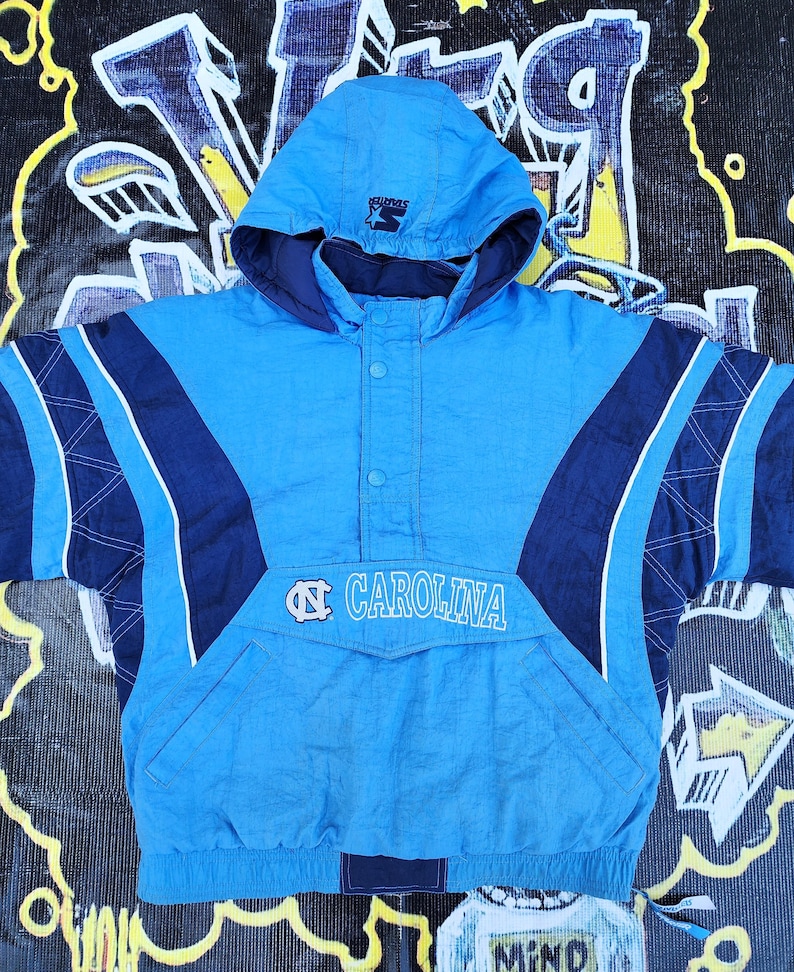 Puede incluir: Chaqueta con capucha azul claro y azul marino con la palabra "CAROLINA" y un logotipo en la parte delantera. La chaqueta tiene un bolsillo delantero y un logotipo de Starter en la capucha. La chaqueta est&aacute; hecha de un material brillante.