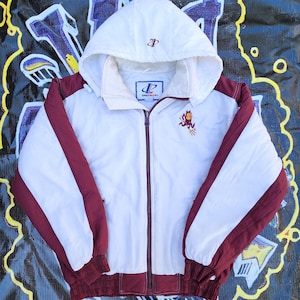 May include: White and maroon zip-up jacket with a hood. The jacket features white panels on the front and sleeves, with maroon accents on the shoulders, sleeves, and hem. The jacket has a small embroidered logo on the chest and hood. The jacket is made of a lightweight material.