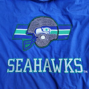May include: A blue jacket featuring the Seattle Seahawks logo. The logo includes a gray football helmet with a green and blue stripe design. The word "SEAHAWKS" is embroidered in white with green accents. The letters "NFL" are embroidered in red.