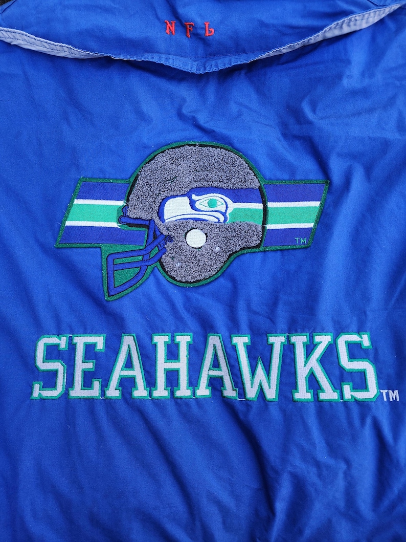May include: A blue jacket featuring the Seattle Seahawks logo. The logo includes a gray football helmet with a green and blue stripe design. The word "SEAHAWKS" is embroidered in white with green accents. The letters "NFL" are embroidered in red.