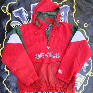 May include: Red, green, and white Devils hooded jacket with a half-zip closure. The jacket features the word "DEVILS" across the chest and a Starter logo. The hood is lined with green fabric.