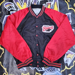 May include: A black and red Tampa Bay Bandits satin jacket. The jacket has red sleeves and a black body with a button-up front. The team logo is on the left chest. The collar and cuffs have red and black stripes.