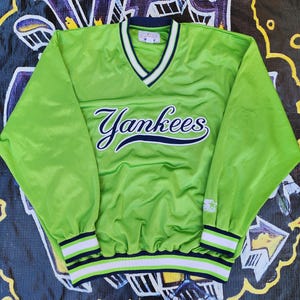 May include: A lime green Yankees baseball jacket with a V-neck collar and long sleeves. The word "Yankees" is written in navy blue script across the chest. The cuffs and hem feature navy blue and white stripes. The Starter logo is visible on the sleeve.