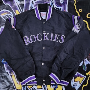 May include: Black satin baseball jacket with purple and white striped trim. The jacket has a large "ROCKIES" embroidered on the front and a "Colorado Rockies" patch on the sleeve.